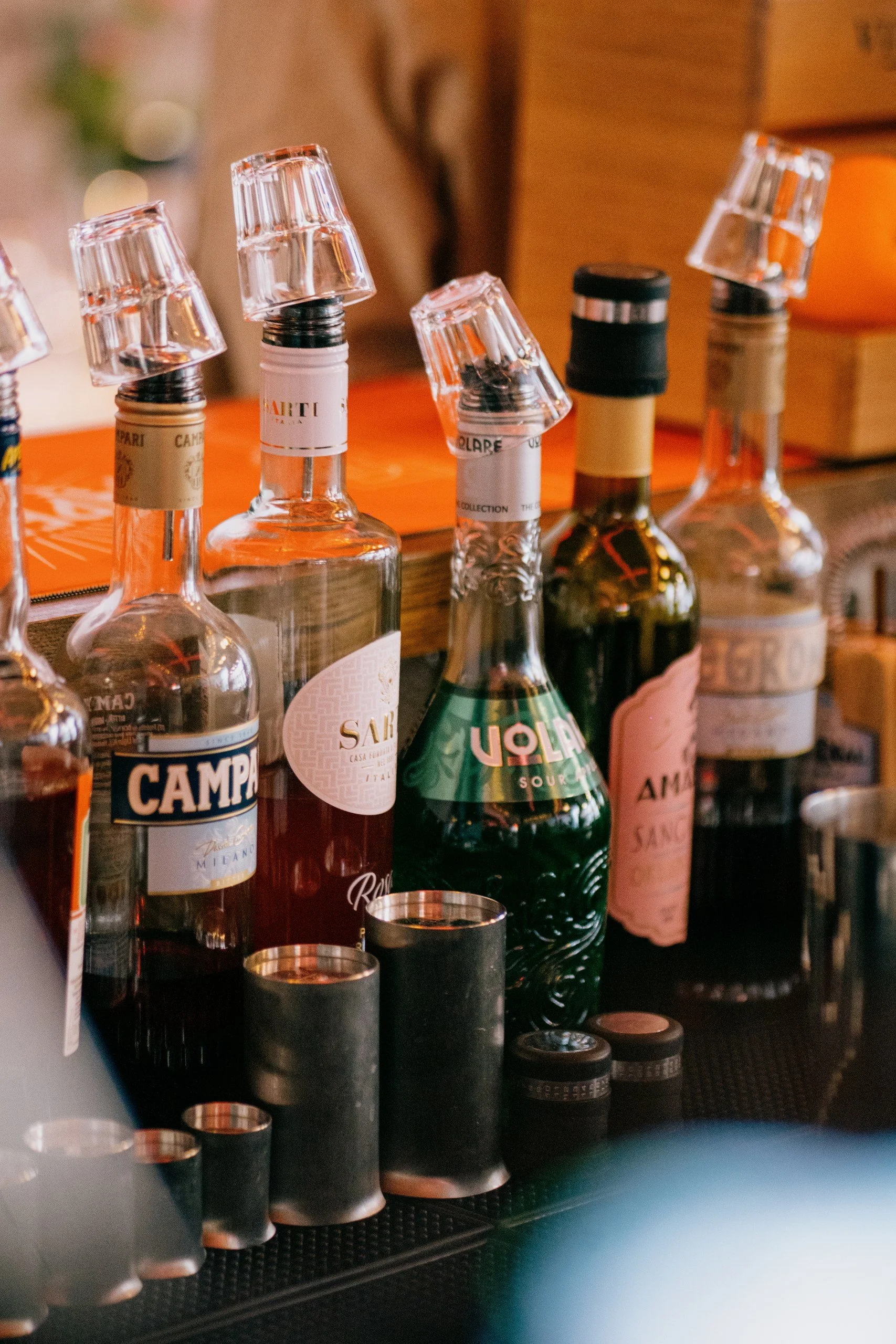 Several bottles of alcohol, including Campari, Sarti, and Amaro, are upside down with shot glasses placed on top of their necks, arranged on a bar counter.