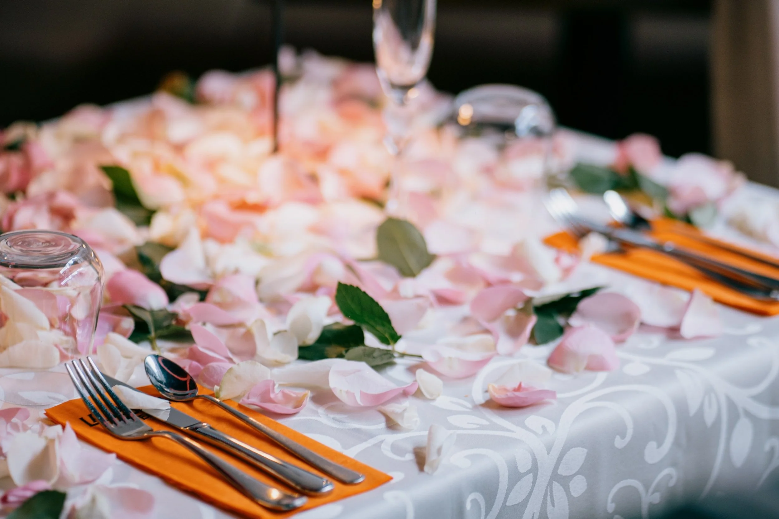 A table decorated with pink and white rose petals, green leaves, clear glassware, utensils, and orange napkins on a white tablecloth.