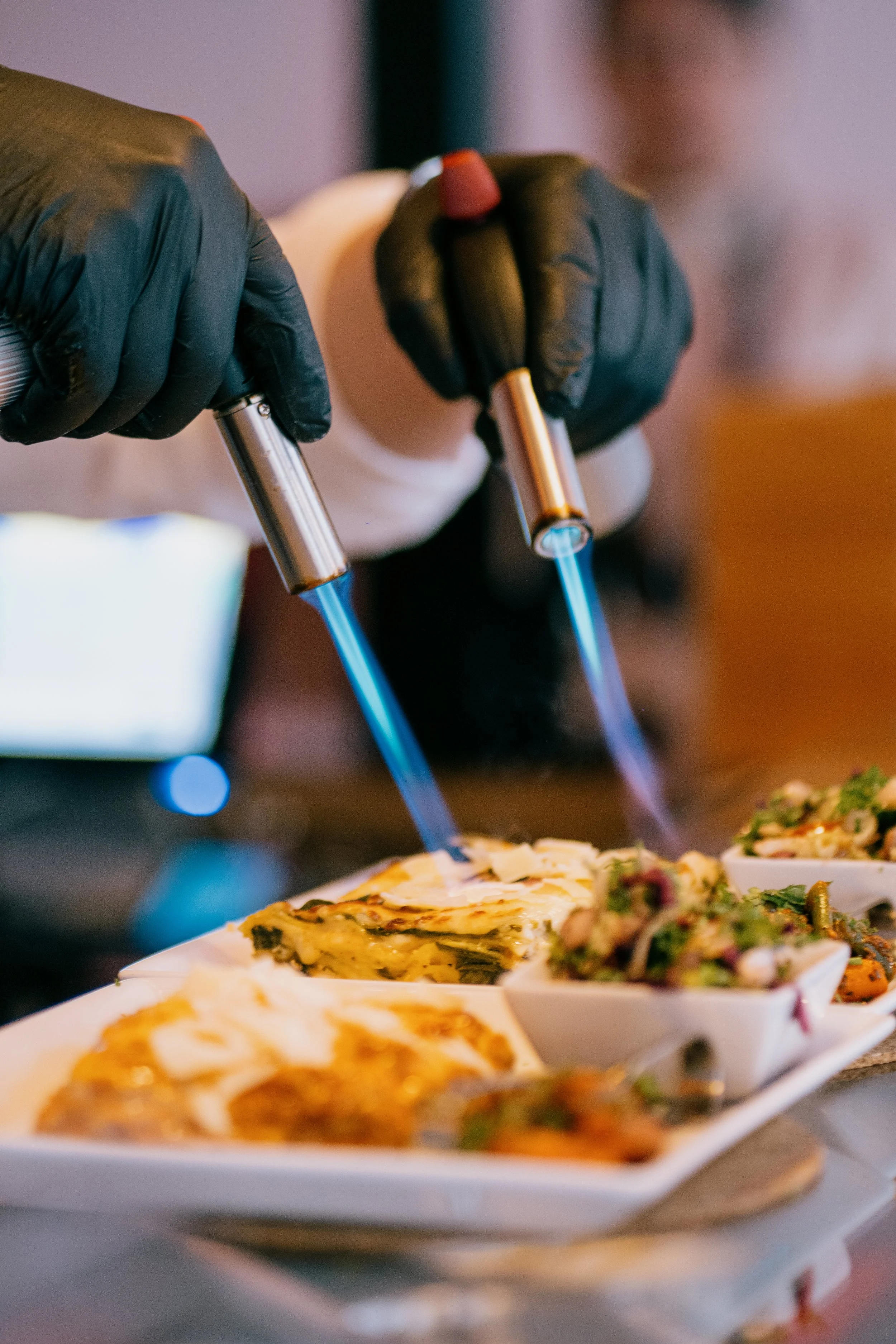 Chef using culinary blowtorches to caramelize or sear food on a white platter with various dishes.