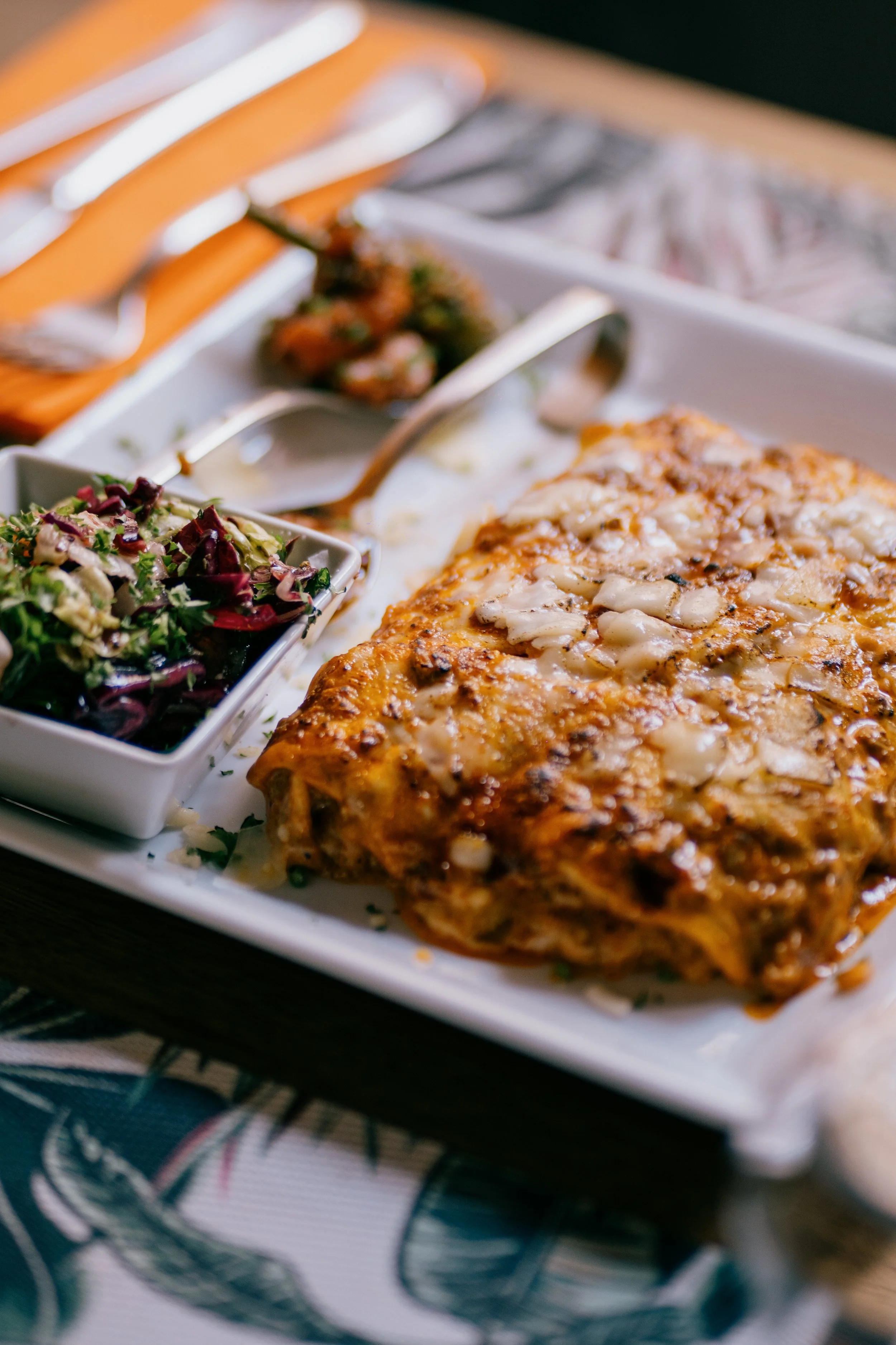 A plate with baked fish topped with onions, a side of mixed greens salad, and remaining food and utensils in the background on a dining table.