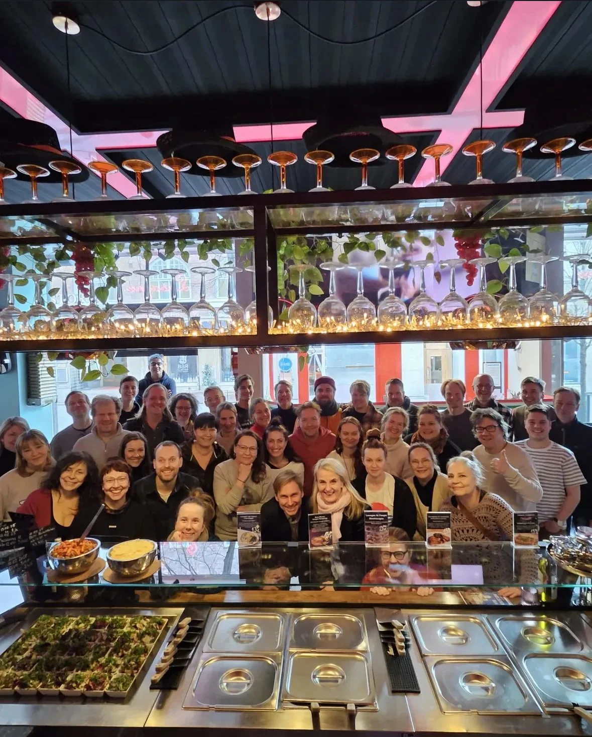 A large group of people smiling in a restaurant with food buffet in the foreground, hanging wine glasses above, and a window letting in natural light.