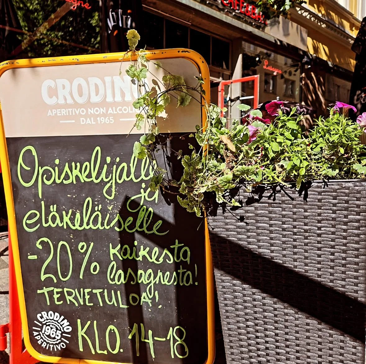 Outdoor restaurant signboard with a potted plant hanging on the side and green handwritten text in Finnish, advertising a 20% discount on lasagna, with a background of street scenes and buildings.