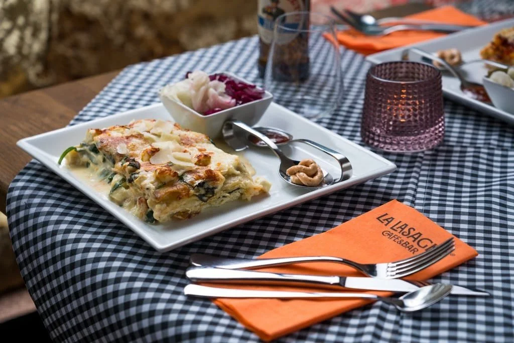 A slice of vegetable and cheese lasagna on a white square plate, with a side of pickled vegetables and a small scoop of nuts, placed on a black-and-white checkered tablecloth at a restaurant.