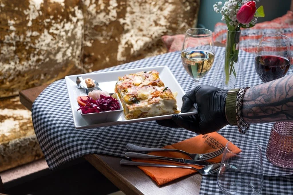A person in black gloves and tattooed arm is serving a slice of vegetable lasagna on a white square plate, accompanied by a small bowl of red cabbage slaw, on a checkered tablecloth. The table has two glasses of white and red wine, a small pink candl
