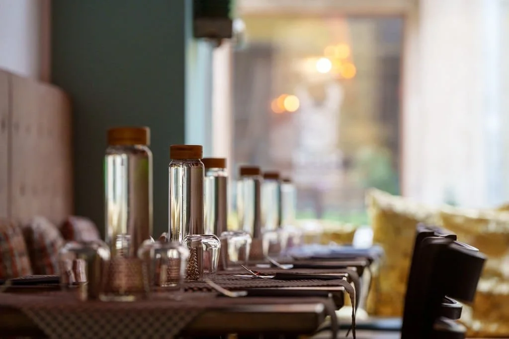 A dining table set with glass water bottles, glasses, and napkins near a window with yellow cushions in the background.