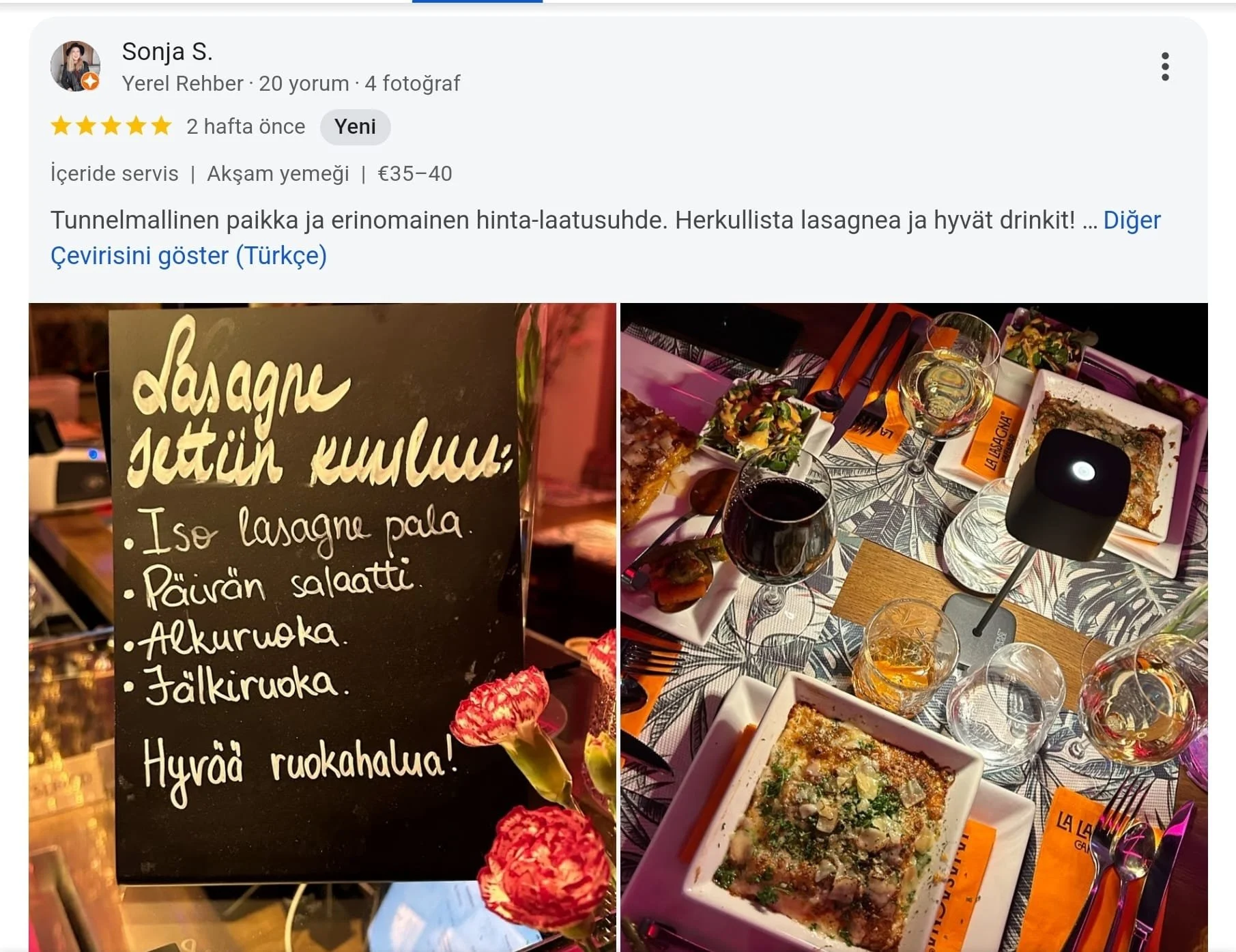 A menu board in Finnish and a table set for a meal at a restaurant, with plates of lasagna, glasses of red and white wine, cutlery, and a black lamp in the center.